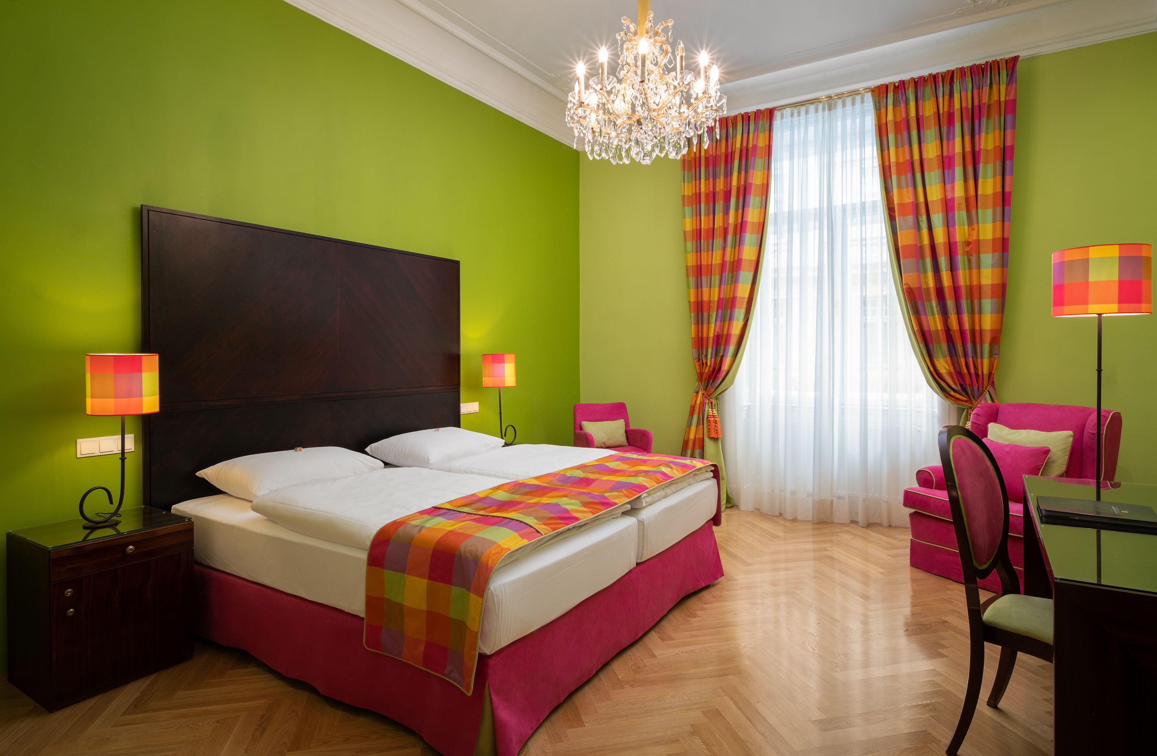 A hotel room with a large bed, a chandelier, a desk with a chair, and a window with curtains.