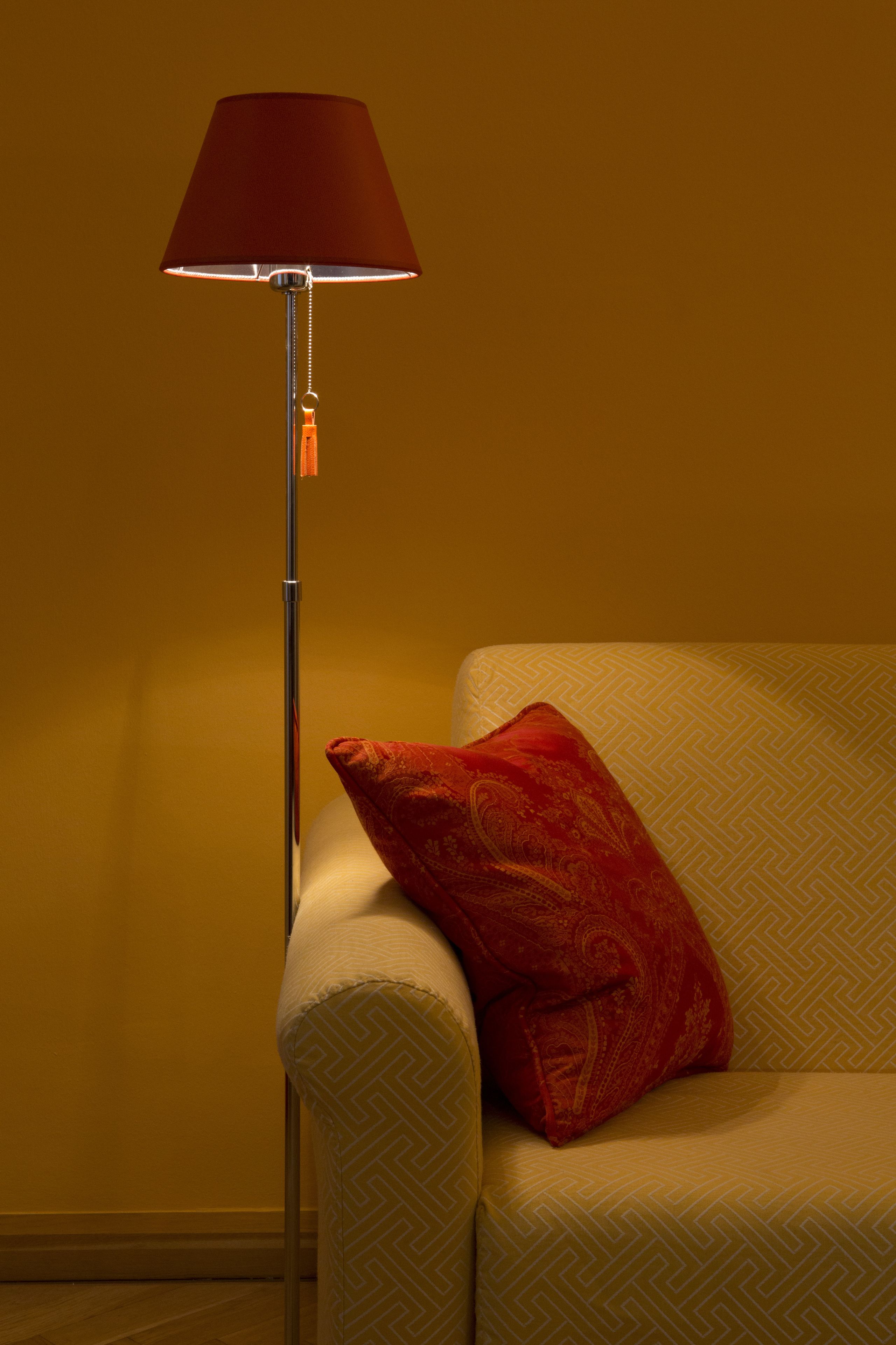A floor lamp with a red shade next to a sofa with a patterned cushion.