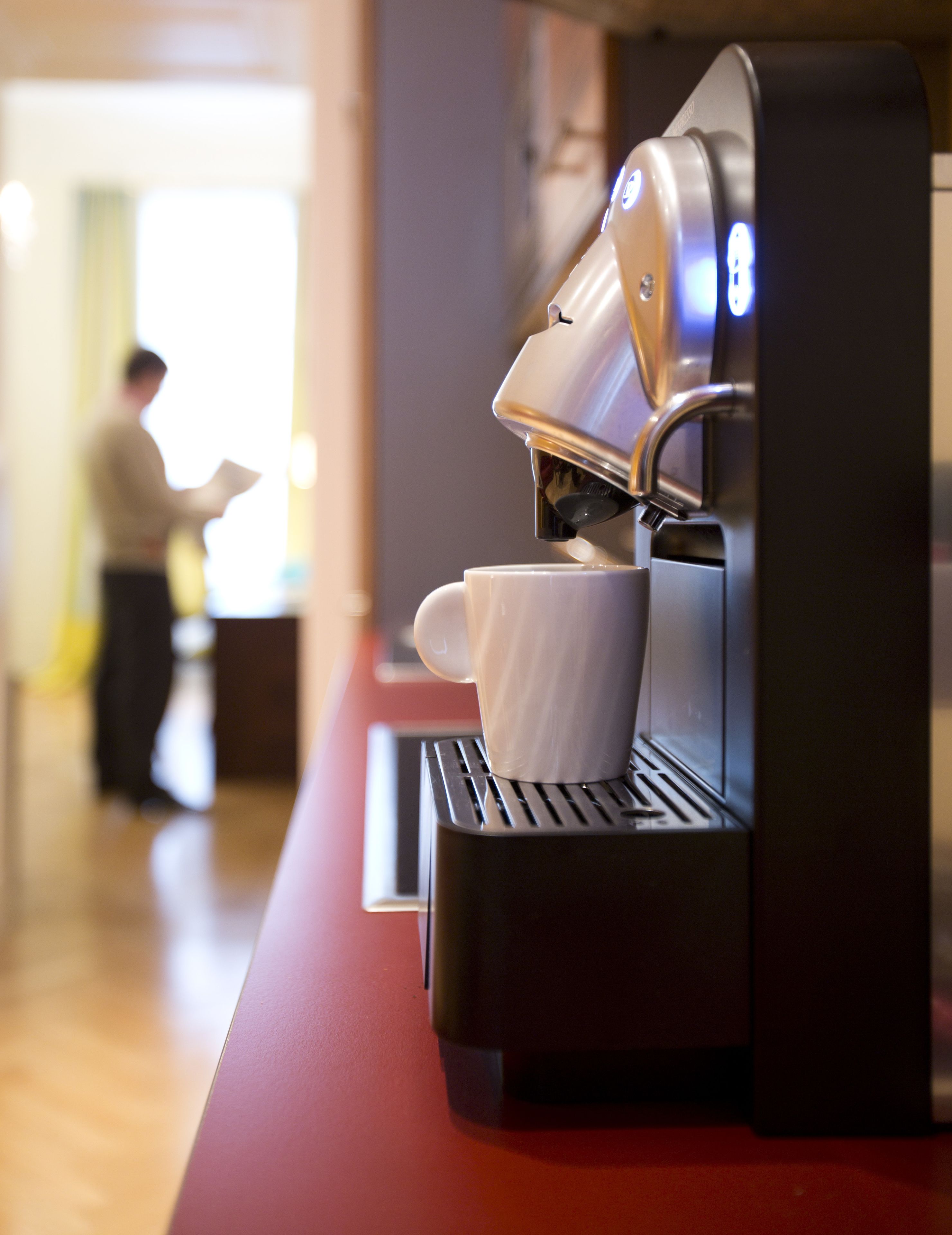 Een koffieapparaat met een kopje eronder, een persoon op de achtergrond en een rode toonbank.
