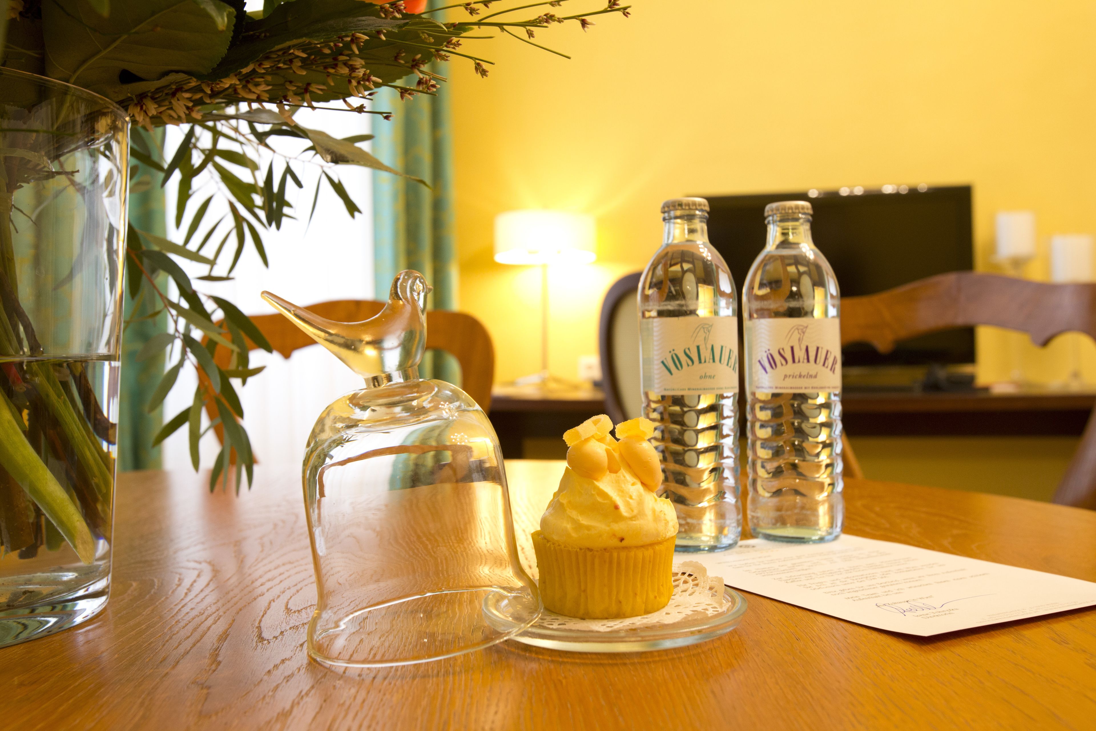 A wooden table with a clear glass decanter, a glass pitcher, and two bottles labeled "Vaslauer" with a cupcake on a doily.
