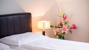 A neatly made bed with white linens, a dark headboard, a bedside lamp, and a vase with pink flowers. - Hotel Pension Dormium (Vienna)