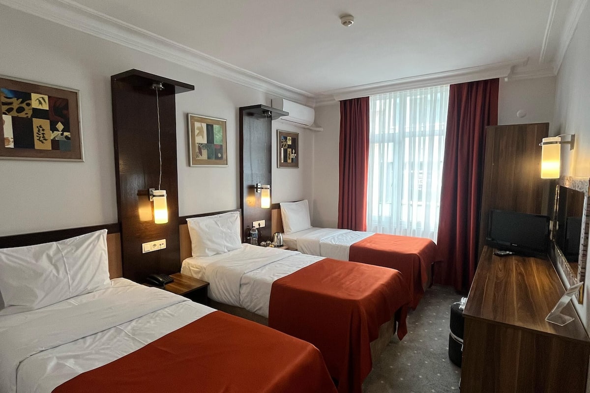 A hotel room with three single beds, a wooden desk, a television, and artwork on the walls.