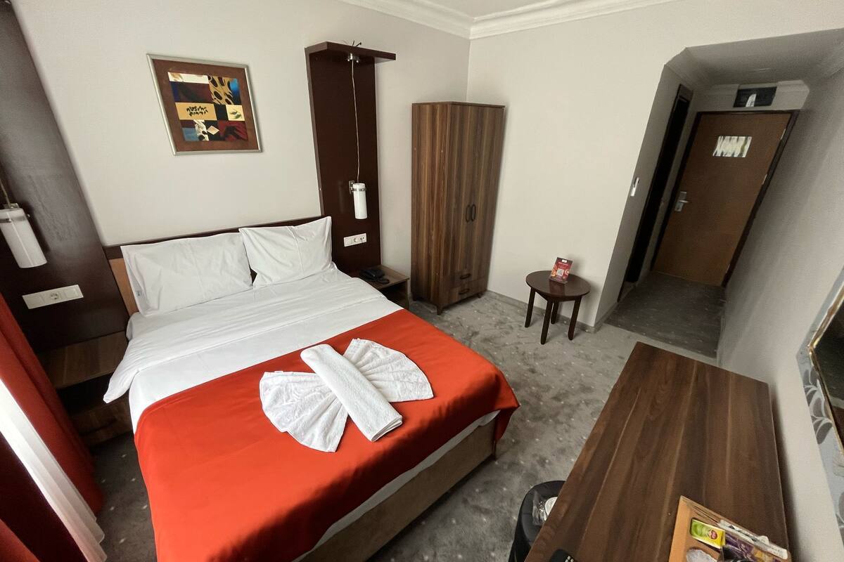 A hotel room with a bed, a wooden wardrobe, a small table with a remote control, and a framed picture on the wall.