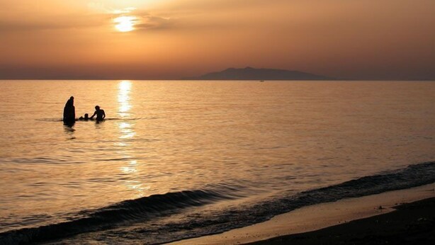 Un atardecer sobre un mar tranquilo, con la silueta de una persona y un niño en el agua.