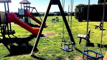 Children can play on the swing set and slide in this playground area, surrounded by a grassy field and a pavilion for shade.