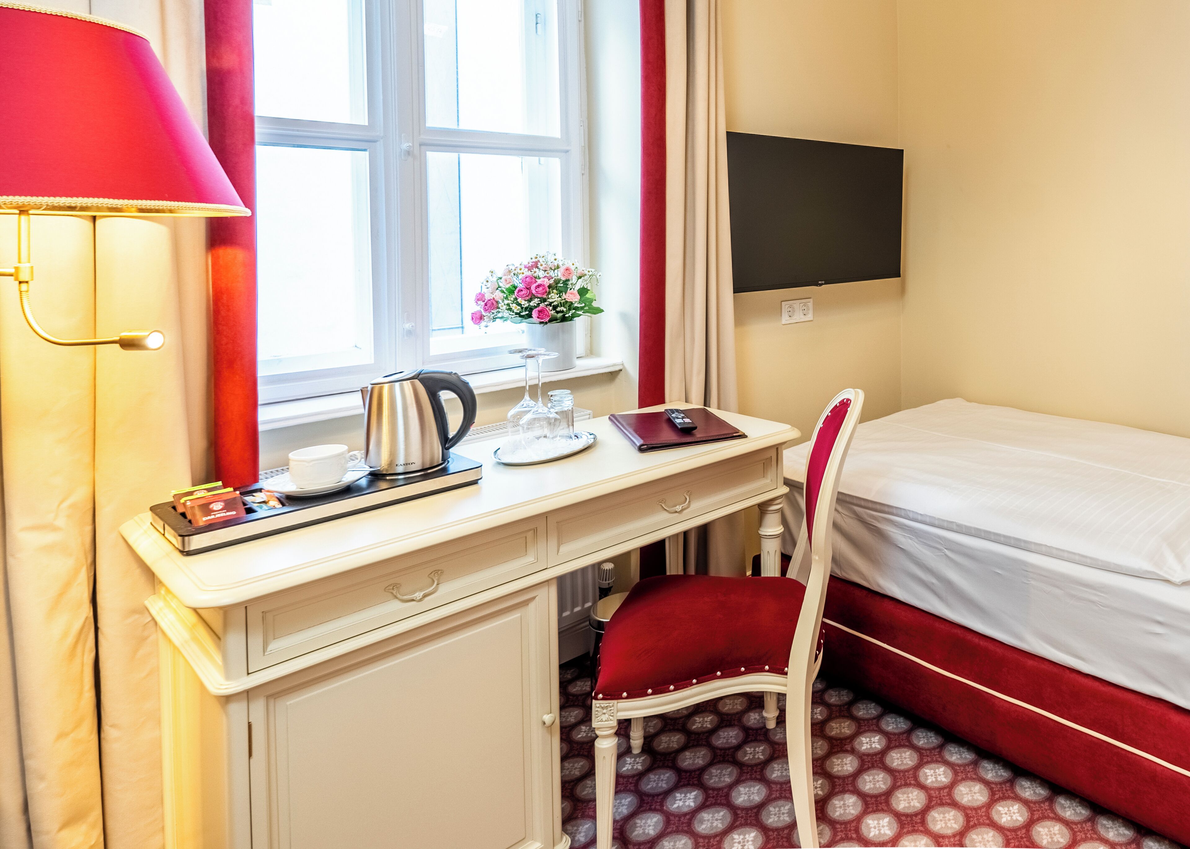Ein Hotelzimmer mit Schreibtisch, Stuhl, Bett, Fernseher, einem Fenster mit Vorhängen, einem Wasserkocher, einer Vase mit Blumen und einer Lampe.