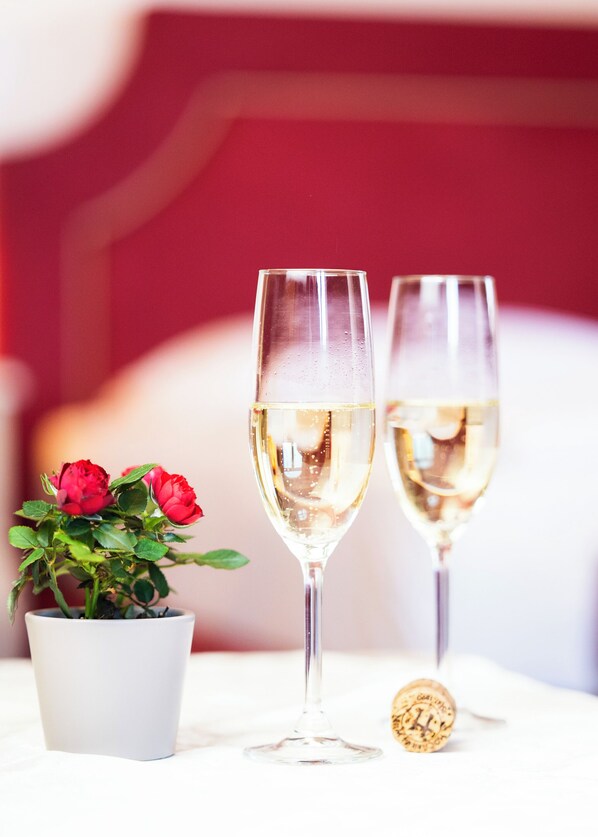Deux flûtes à champagne, dont l’une est fermée par un bouchon, sont posées sur une table blanche. On aperçoit également un pot de fleurs contenant des roses rouges, et un fond à motifs rouges et blancs.