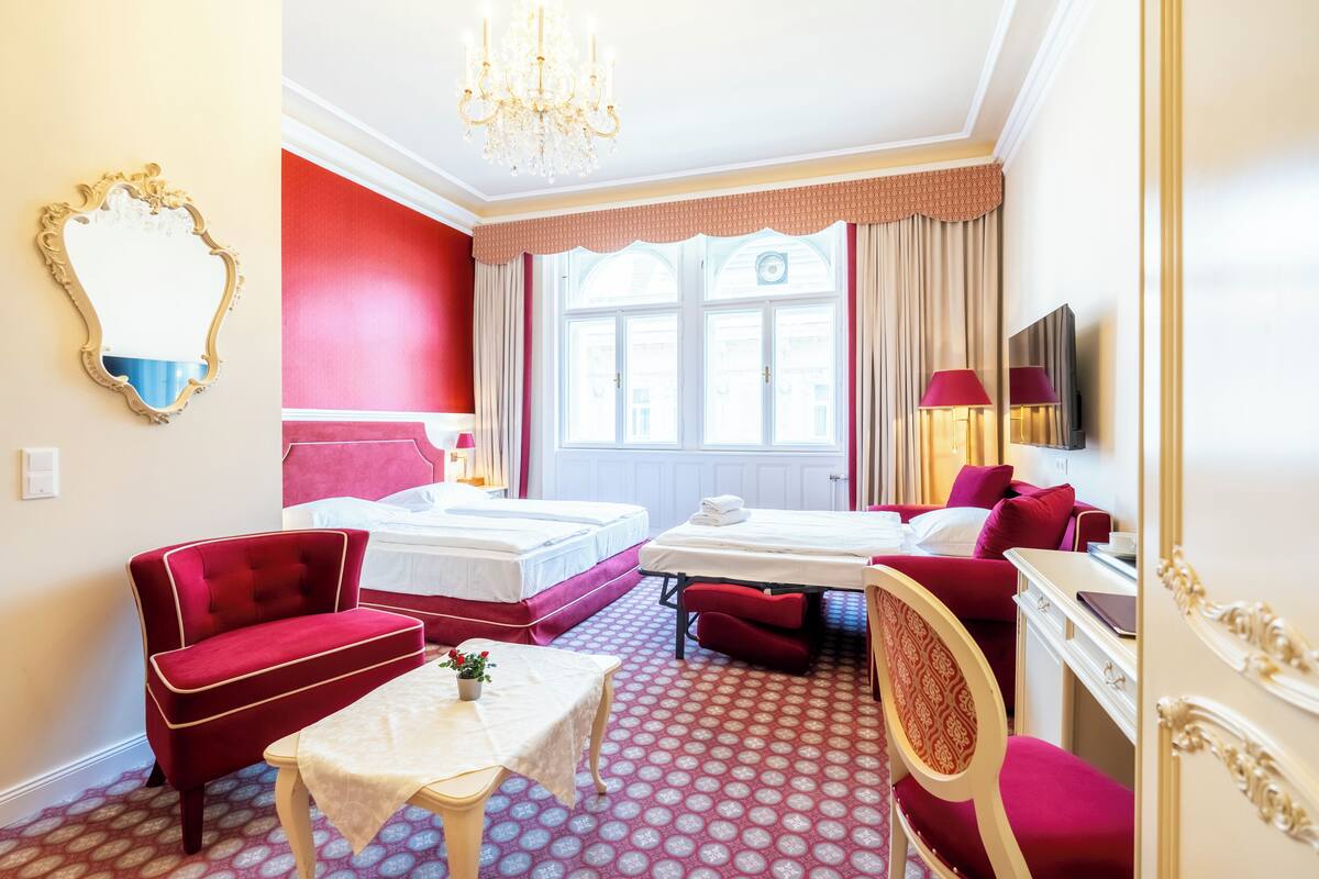 A hotel room with two beds, a red sofa, a small table with a plant, a TV, and a chandelier.