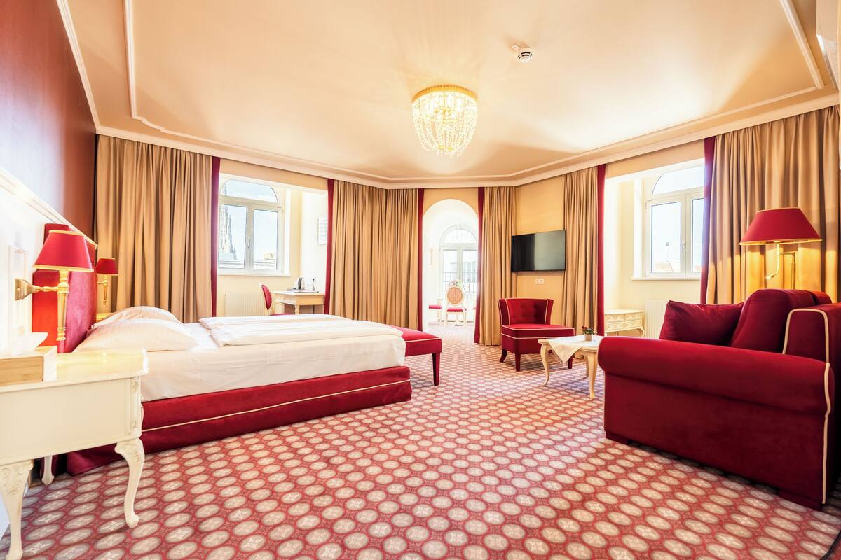 A hotel room with a large bed, a red armchair, a TV, and a chandelier.