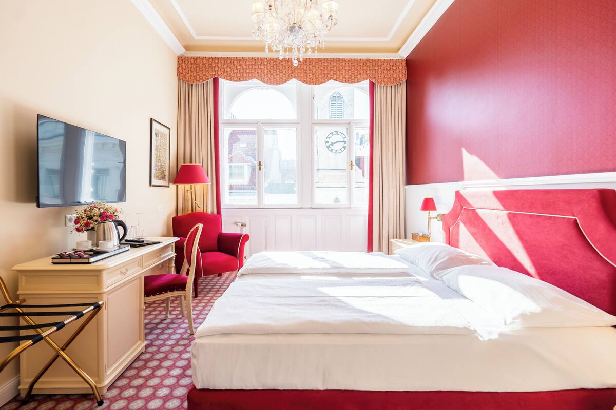 A hotel room with a bed, a desk with a flat-screen TV, a chair, a desk lamp, and a window with curtains.