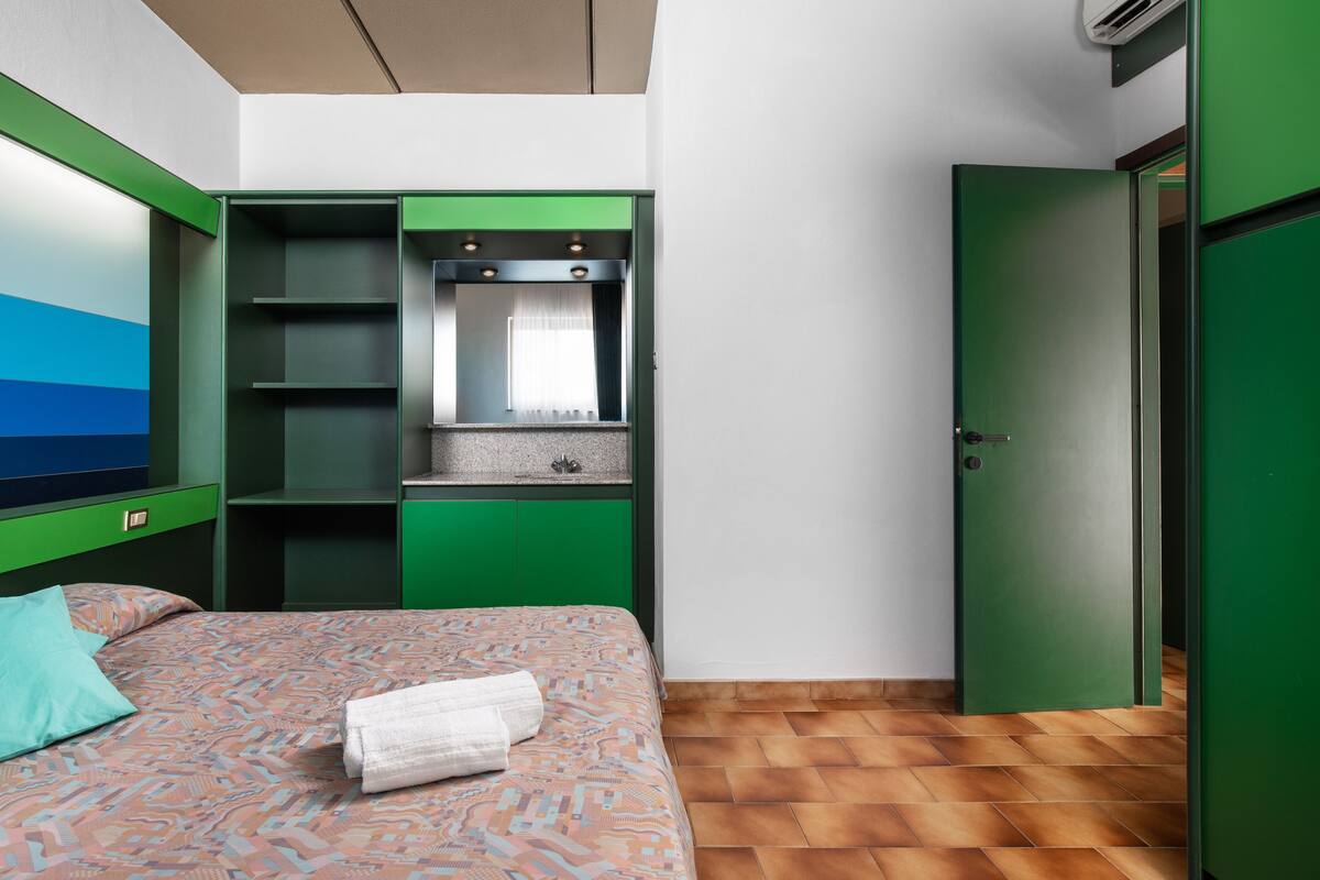 A room with a bed, a green wardrobe, a mirror, and a window with curtains.