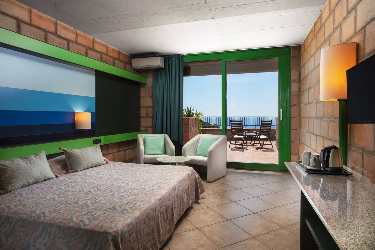 A hotel room with a bed, two armchairs, a small table, a balcony with a view, and a TV.