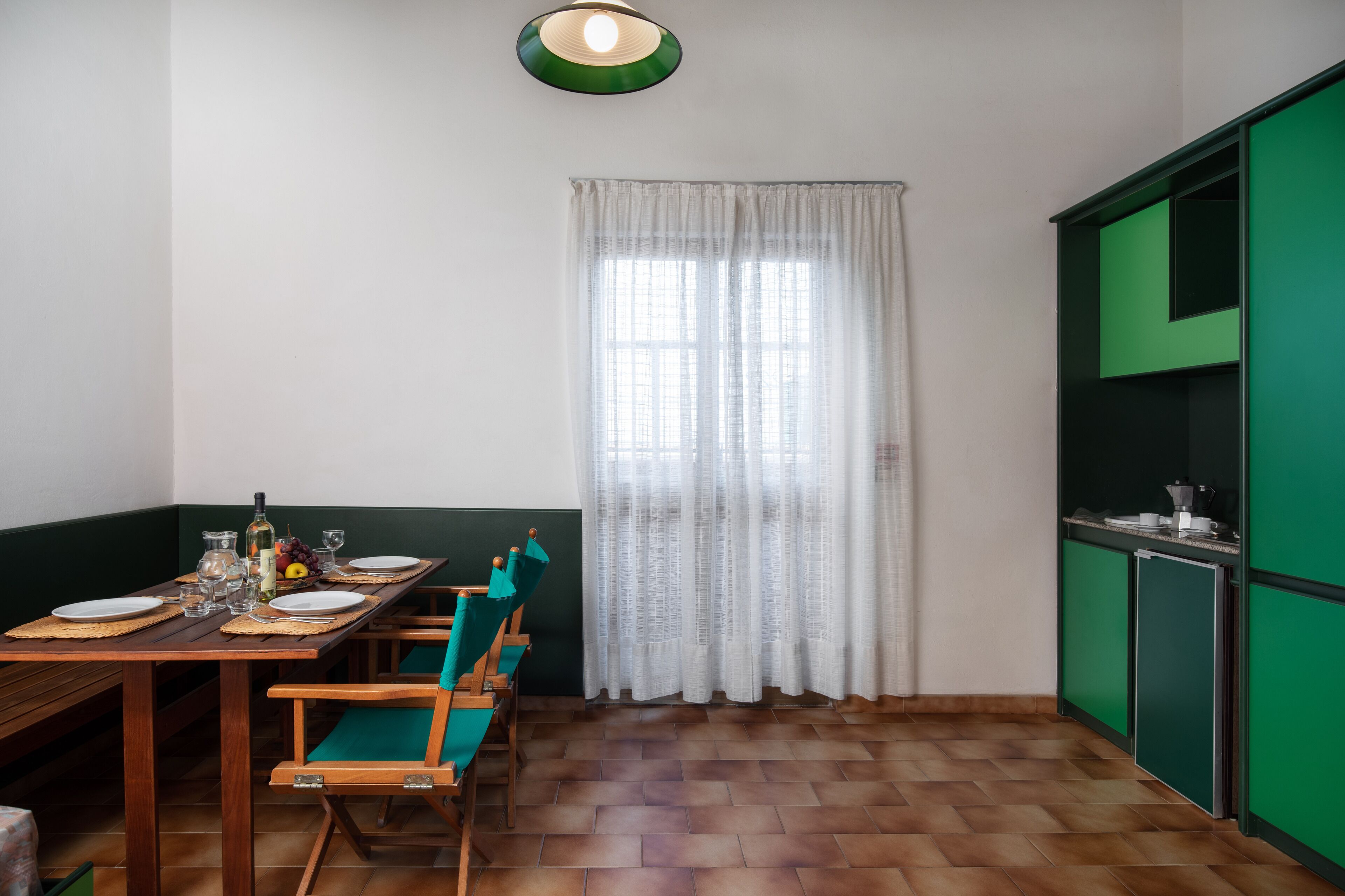 A dining area with a wooden table set for four, chairs, a window with white curtains, and a green cabinet.