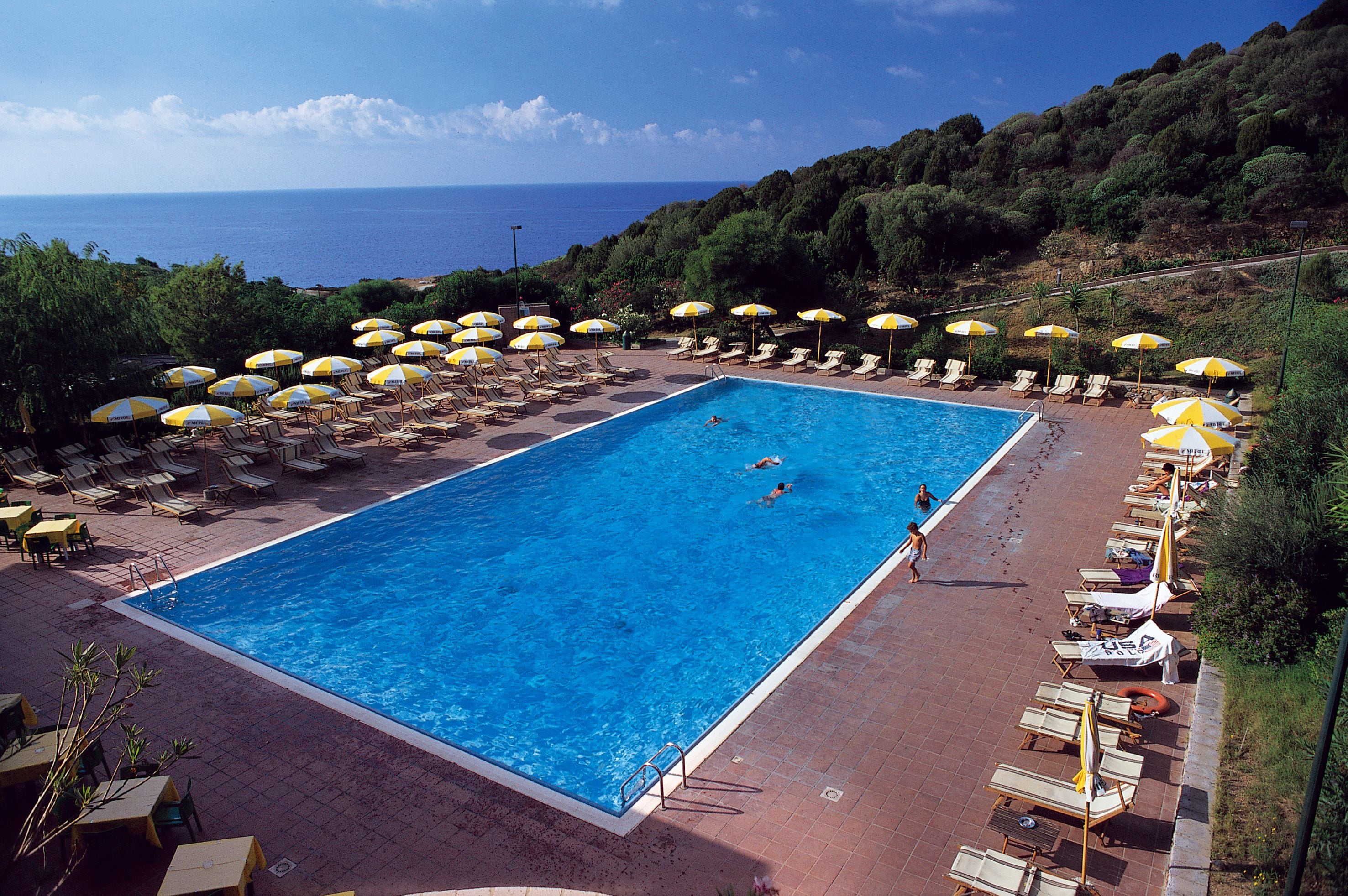 Ein großes rechteckiges Schwimmbecken mit klarem, blauem Wasser, umgeben von Liegestühlen und gelben Sonnenschirmen, mit Blick auf Meer und Hügel im Hintergrund.