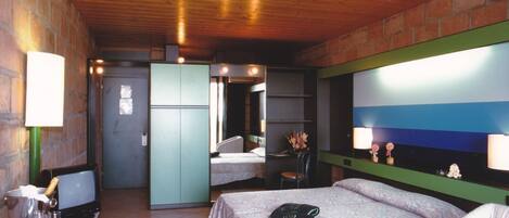 A hotel room with a bed, a desk with a lamp, a television, and a brick wall.
