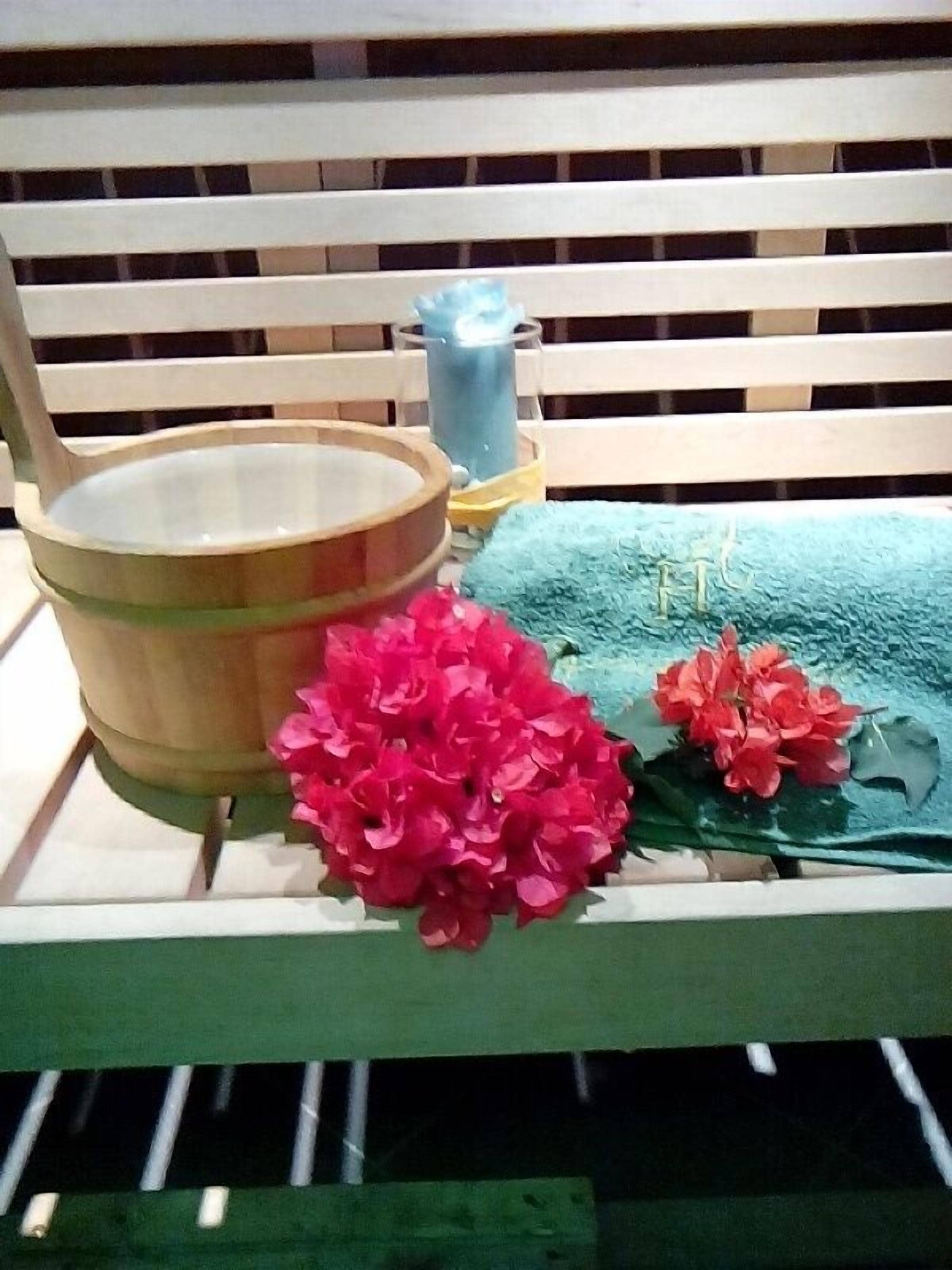 A wooden bench with a metal bucket, a blue container, and red flowers.