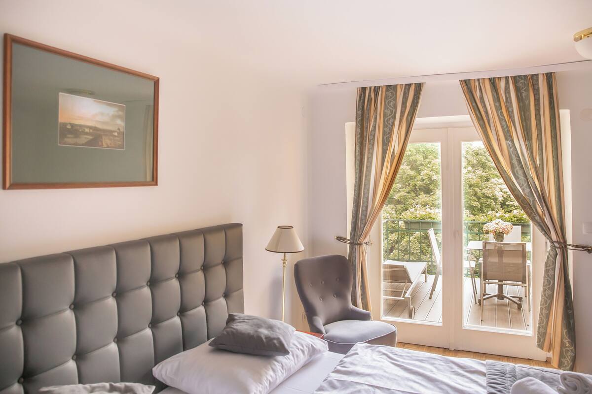 Ein Schlafzimmer mit einem gesteppten Kopfteil, einem Nachttisch, einem Sessel und Blick auf einen Balkon mit Vorhängen.