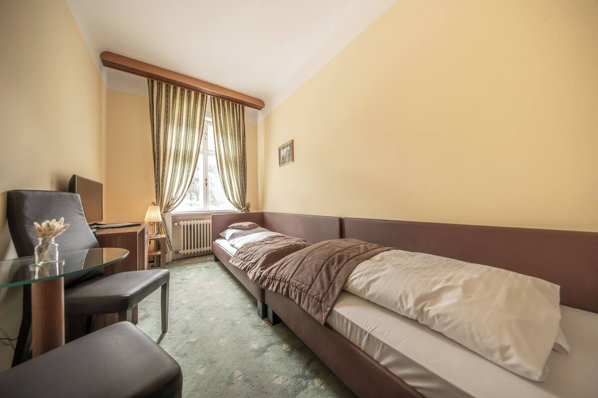 Ein Hotelzimmer mit einem Einzelbett, einem kleinen Tisch mit Stuhl, einem Schreibtisch und einem Fenster mit Vorhängen.