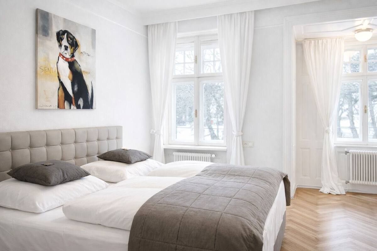 Ein Schlafzimmer mit einem großen Bett, einem Kopfteil, zwei Fenstern mit weißen Vorhängen und einem Hundebild an der Wand.