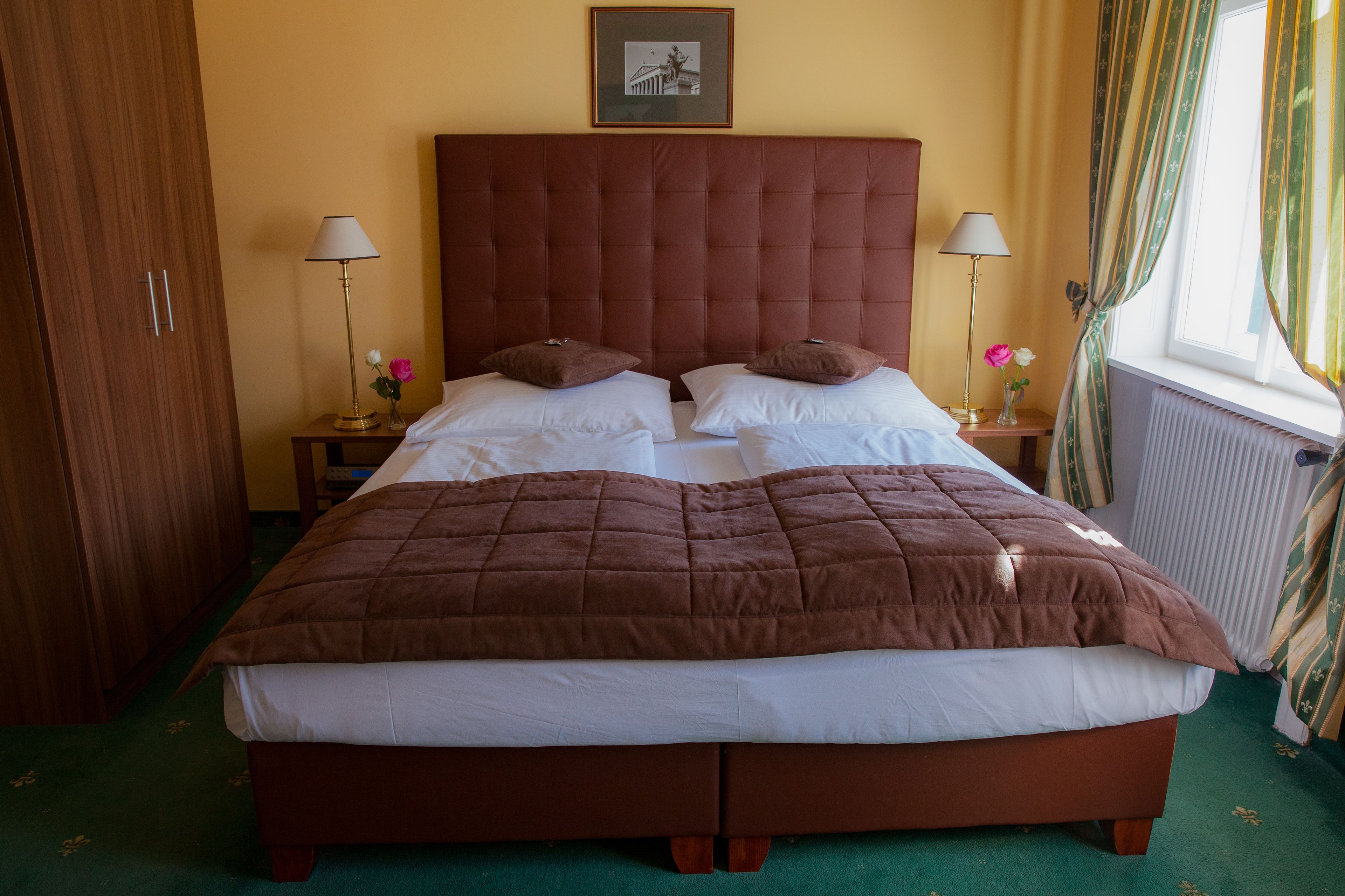 Un lit double avec une couette marron, deux tables de chevet avec des lampes, un tableau encadré au-dessus du lit et une fenêtre avec des rideaux.
