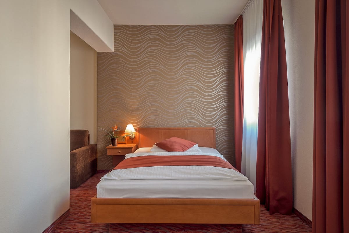 A hotel room with a bed, a nightstand with a lamp, a chair, and a patterned wall.