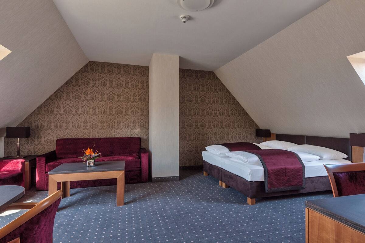 A hotel room with a sloped ceiling, a sofa, a bed with two pillows, a small table with a vase, and patterned wallpaper.