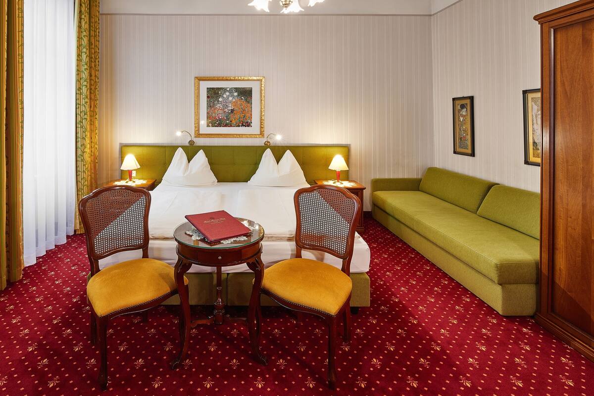 Une chambre d’hôtel avec un lit, deux chaises, une table, un canapé vert et un lustre.