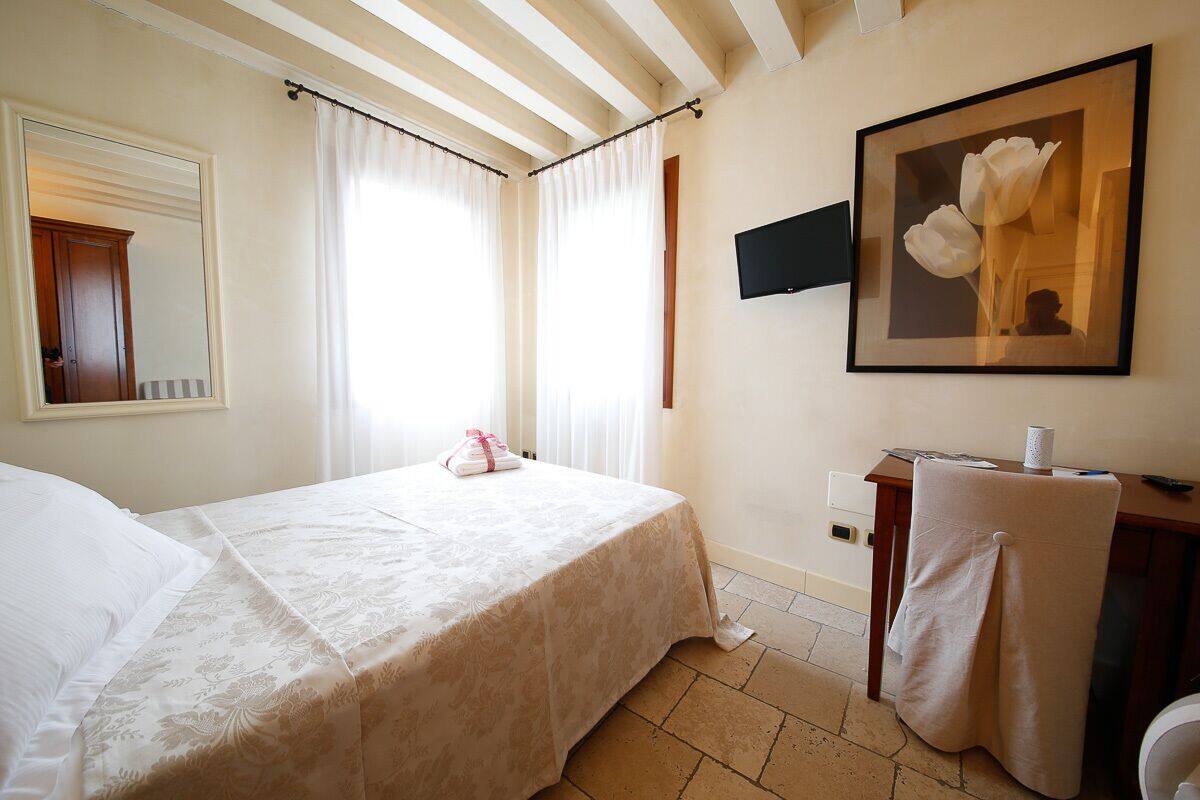 A hotel room with a bed, a desk with a chair, a TV mounted on the wall, and a framed picture of flowers.
