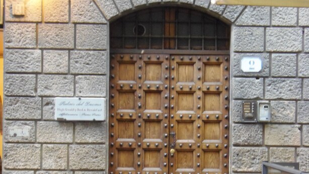 A stone building with a large wooden double door, a sign above the door, and a light fixture on the wall. - Relais del Duomo (Florence)