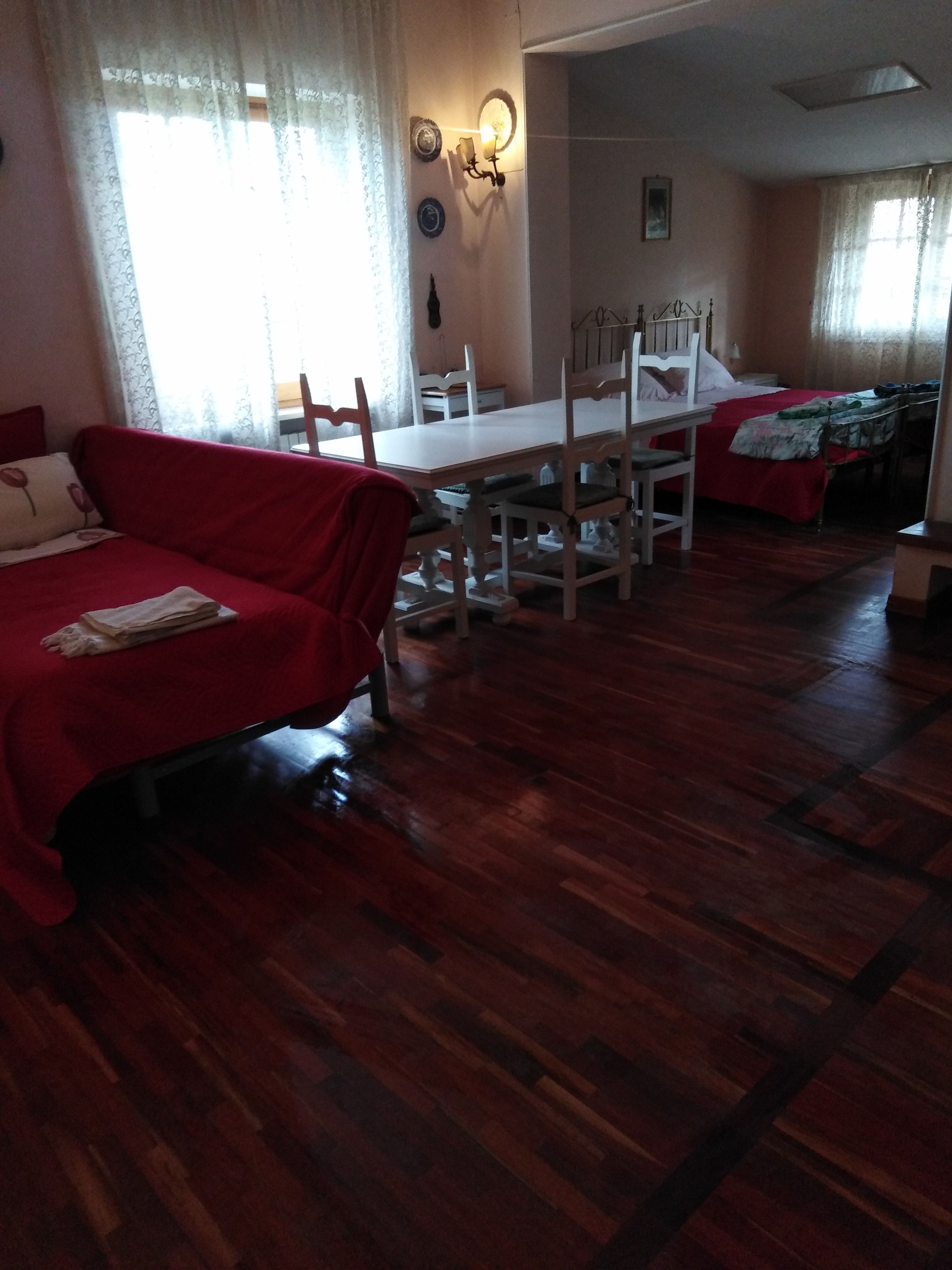 A room with a red sofa, a wooden dining table with chairs, and a bed with a red blanket.