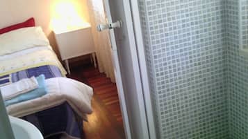 A small, compact room with a bed, a bedside table with a lamp, a white sink, and a shower area with tiled walls.