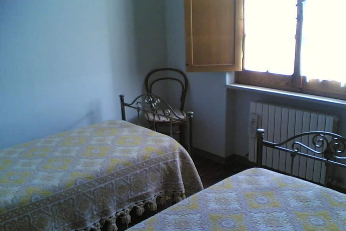 A bedroom with two beds, a wooden chair, a window with a wooden blind, and a radiator.