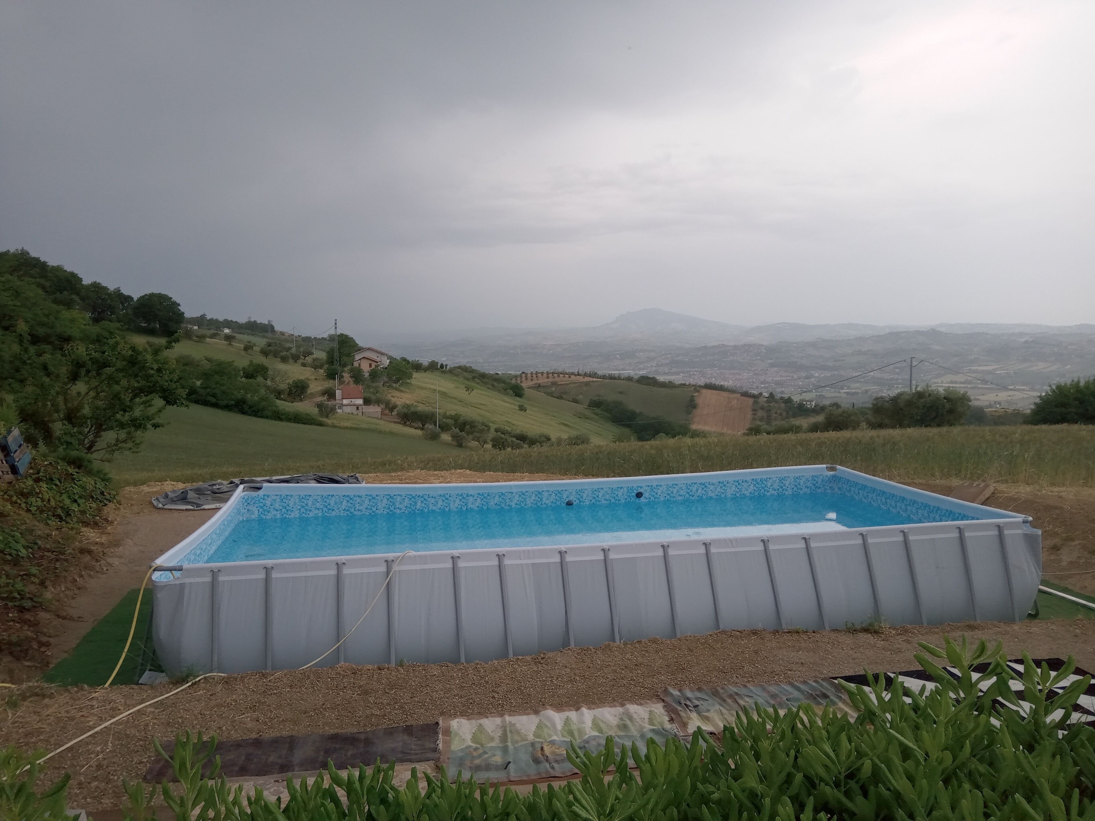 A rectangular inflatable pool with a clear blue interior, surrounded by a grassy area with a view of distant hills and a small building in the background.