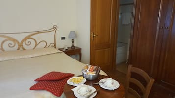 Una cama bien hecha con un cojín, una mesita de noche con una lámpara, una pequeña mesa con cosas para el desayuno y un armario de madera.