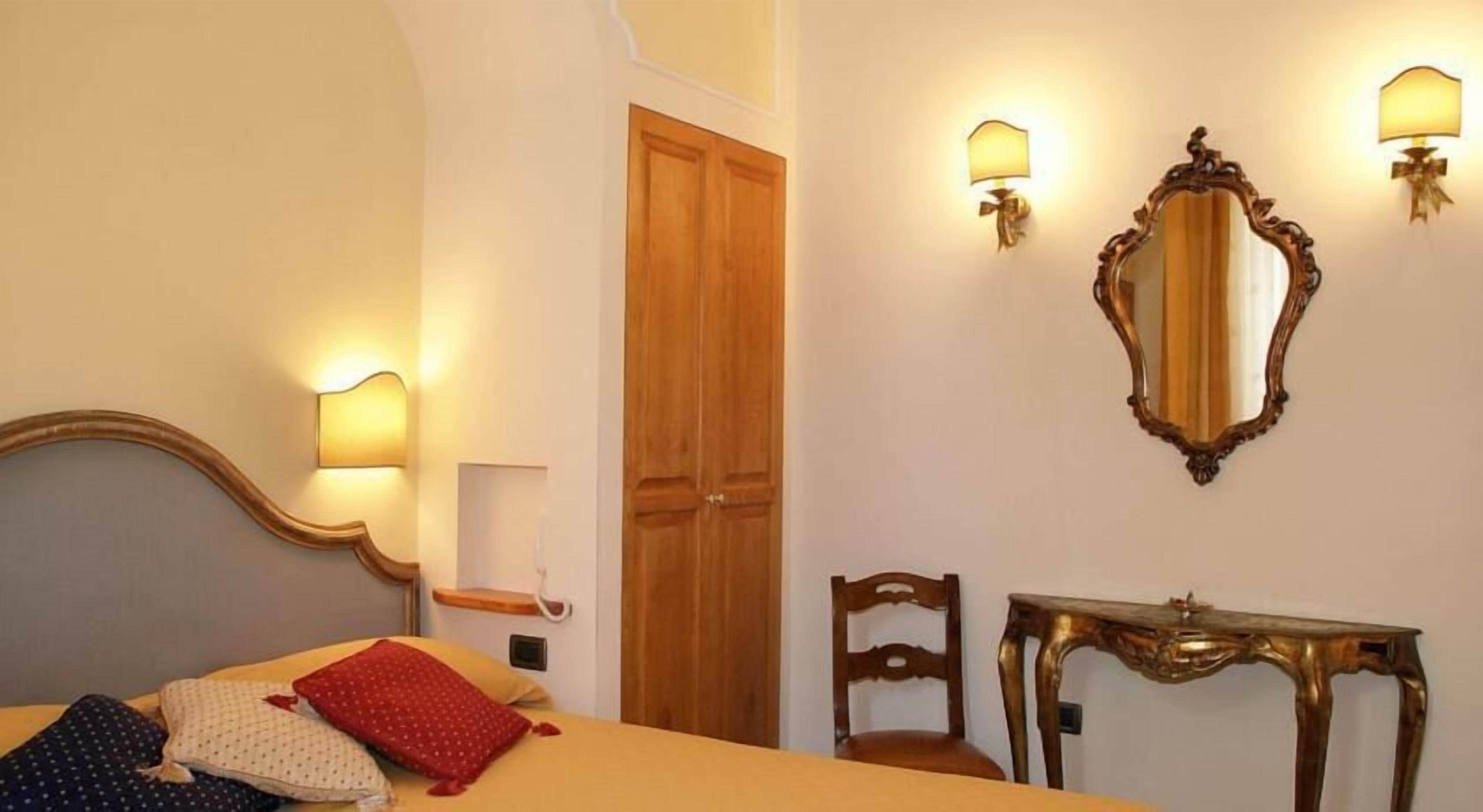 Quarto de hotel com uma cama, uma porta de madeira, uma cadeira, uma pequena mesa e duas luminárias fixadas na parede.
