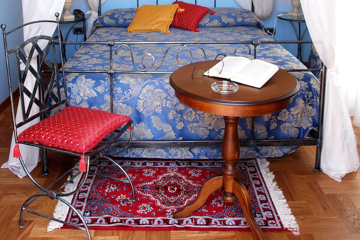 A four-poster canopy bed with a blue floral bedspread, a red cushion, and a wooden nightstand with a book.