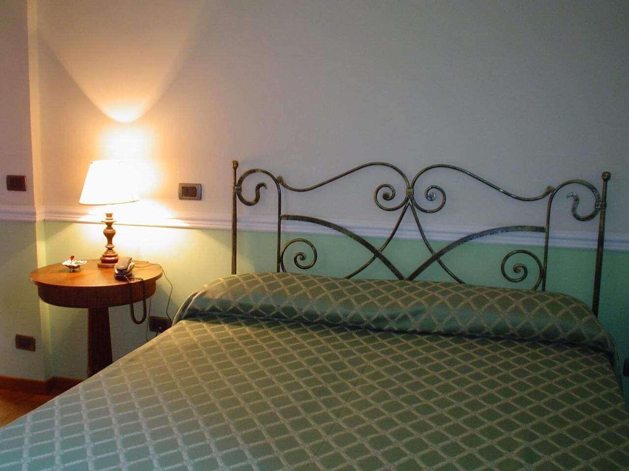 A bed with a metal headboard, a bedside table with a lamp, and a wall-mounted light.