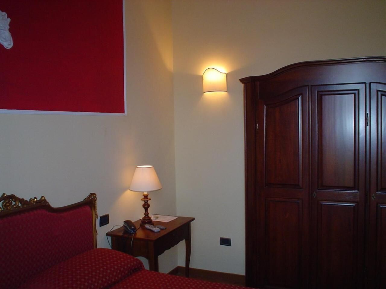 A hotel room with a red bed, a bedside table with a lamp, a wardrobe, and a wall-mounted light fixture.