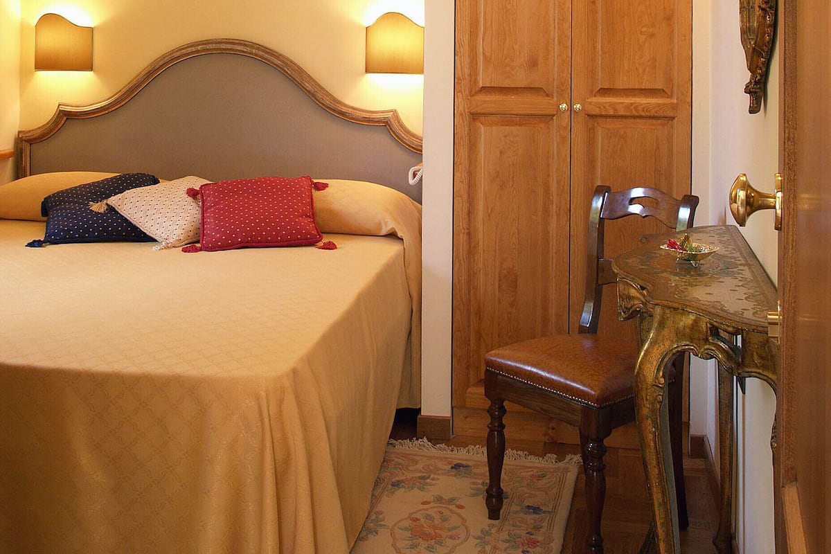 A bedroom with a bed, two bedside lamps, a wooden wardrobe, a chair, a small table, and a framed picture on the wall.