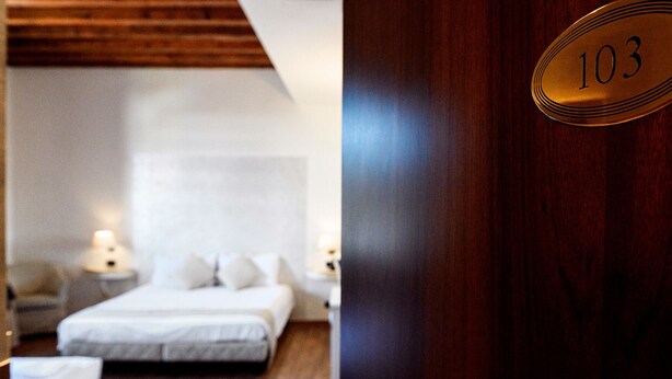 Habitación de hotel con cama, mesitas de noche y una puerta de madera con placa de número.