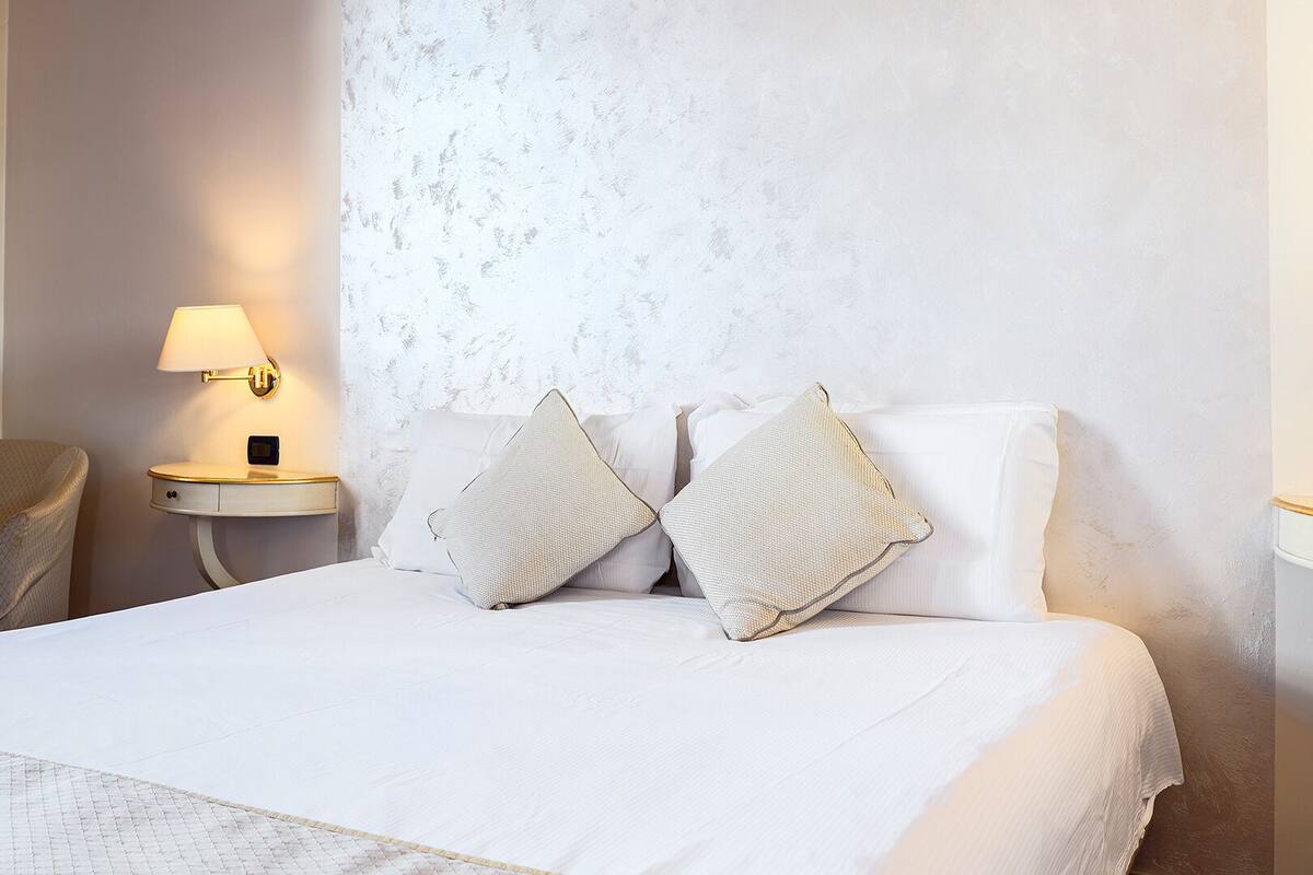 A hotel room with a bed, a nightstand with a lamp, a chair, and a textured wall.