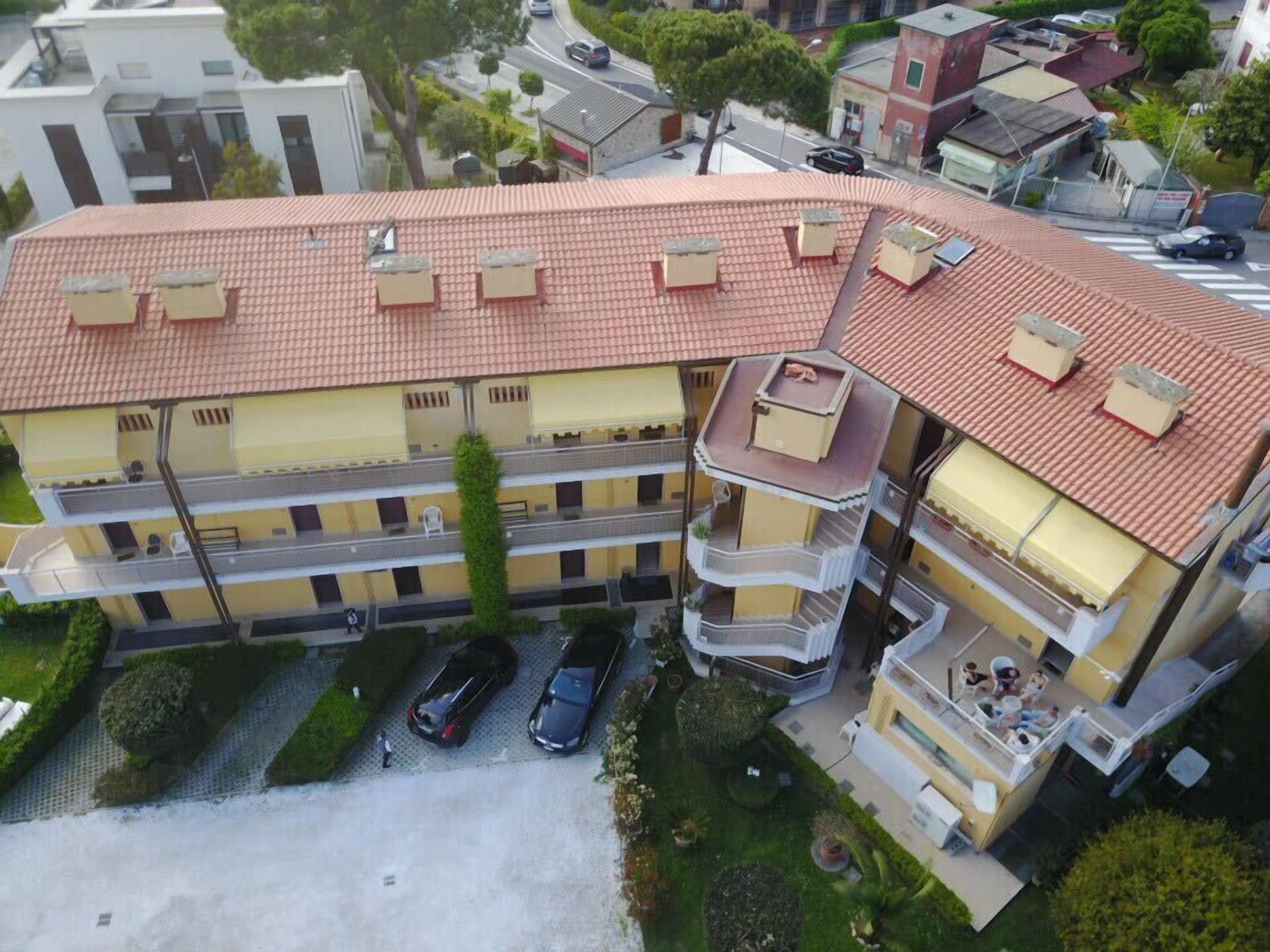 A multi-story residential building with a red-tiled roof, multiple balconies, and a parking area with cars.
