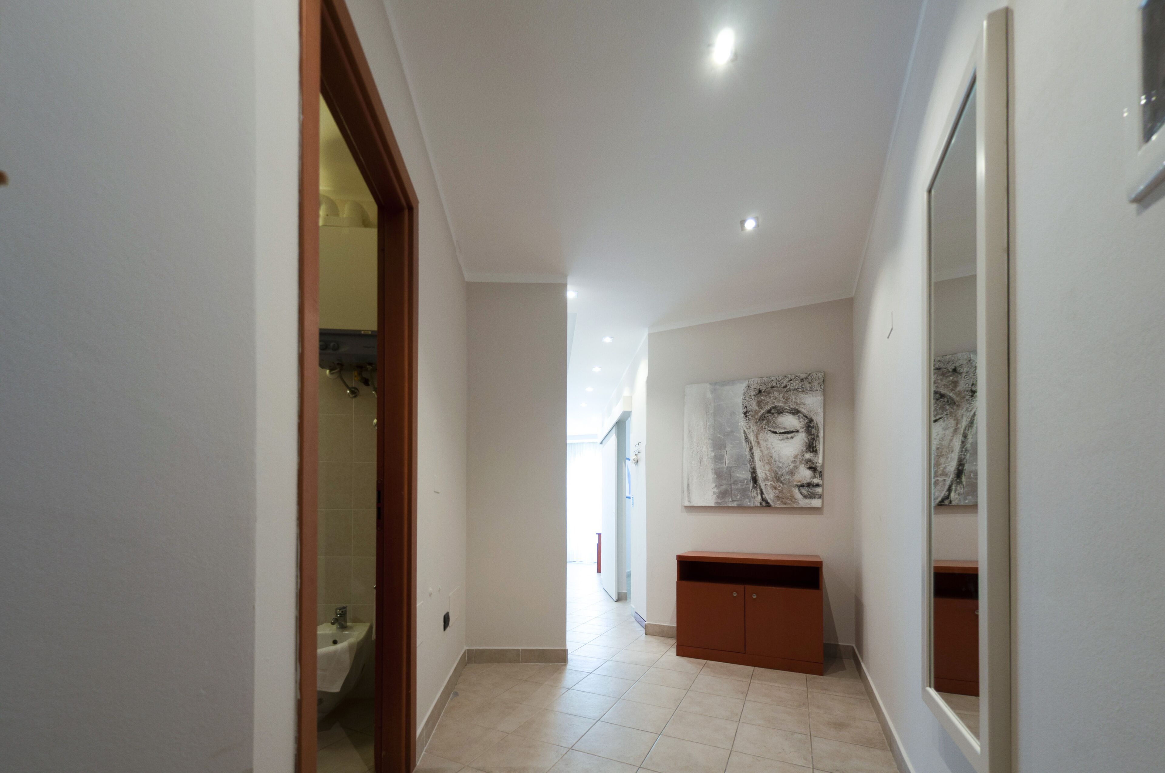 A hallway with a bathroom on the left, a sink, and a cabinet. There are ceiling lights and a large mirror on the right.