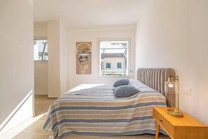 A bedroom with a bed, bedside table, lamp, window, and artwork.