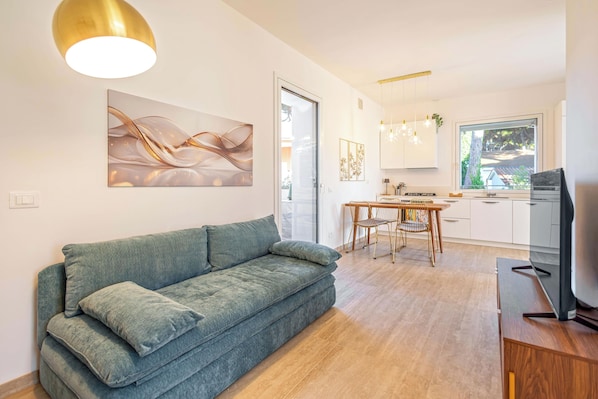 A modern living room with a sofa, a dining area with a table and chairs, a kitchen with cabinets and a window, and a television on a wooden stand.