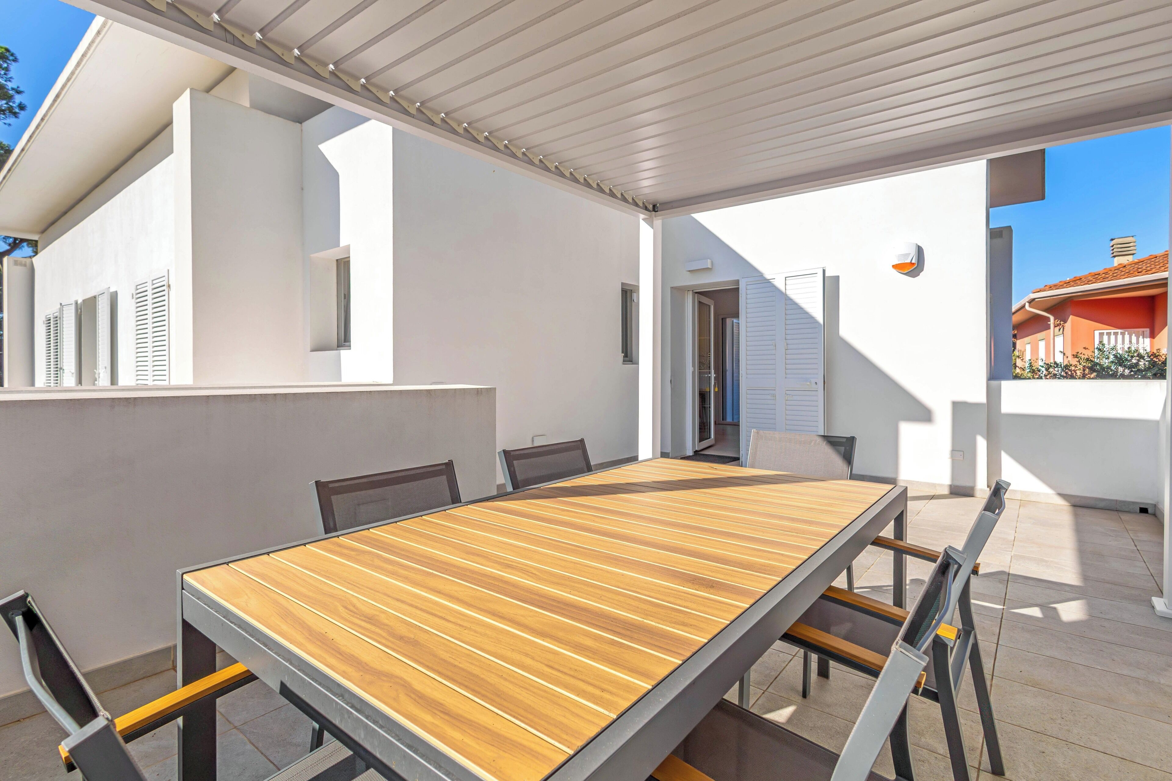 Un espacio exterior techado para comer con una mesa y sillas de madera, paredes blancas y cielo despejado.