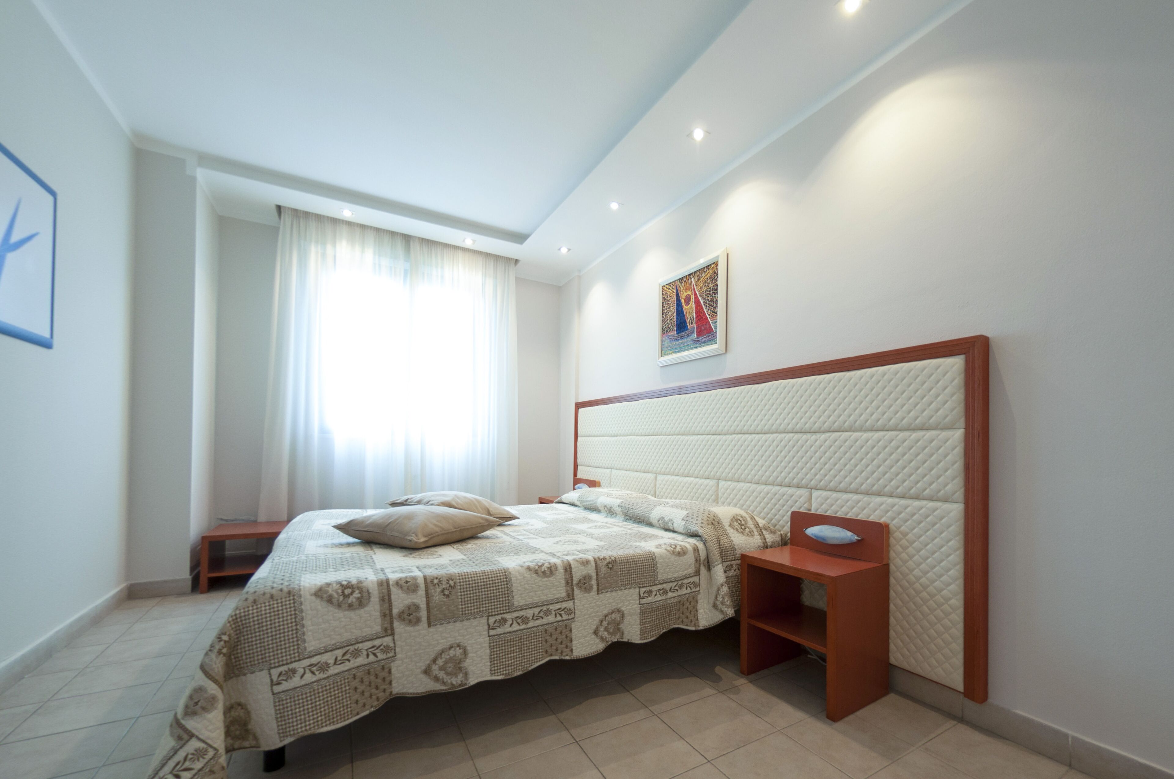 Ein Schlafzimmer mit einem Bett, Nachttischen, einem Fenster mit Vorhängen und einem Wandbild.