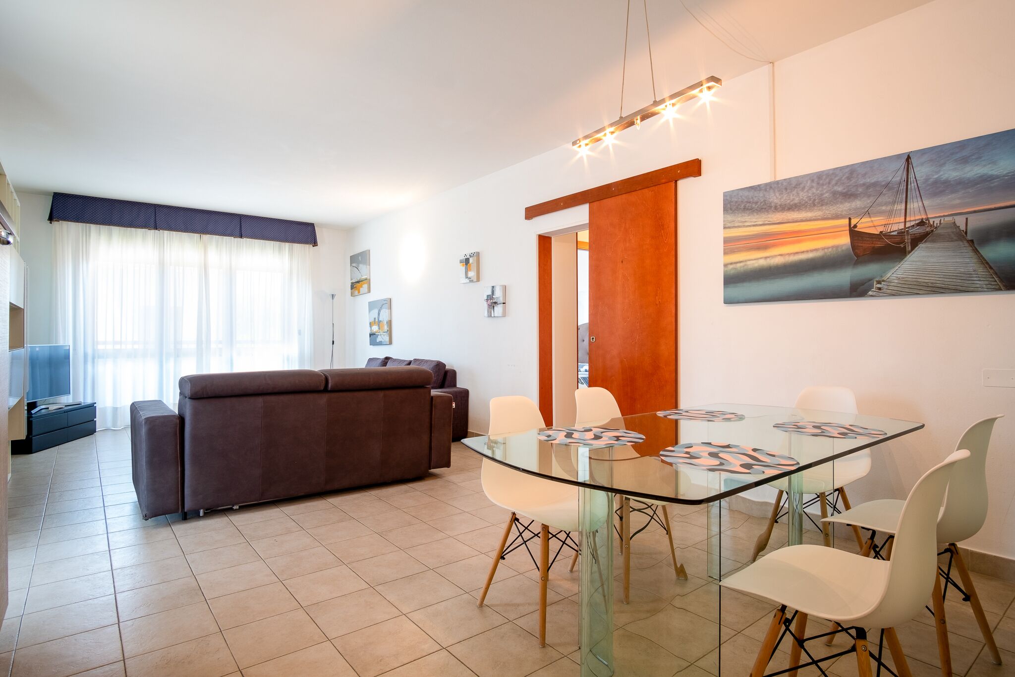 A modern living room with a glass dining table, white chairs, a brown sofa, and a painting of a sunset on the wall.