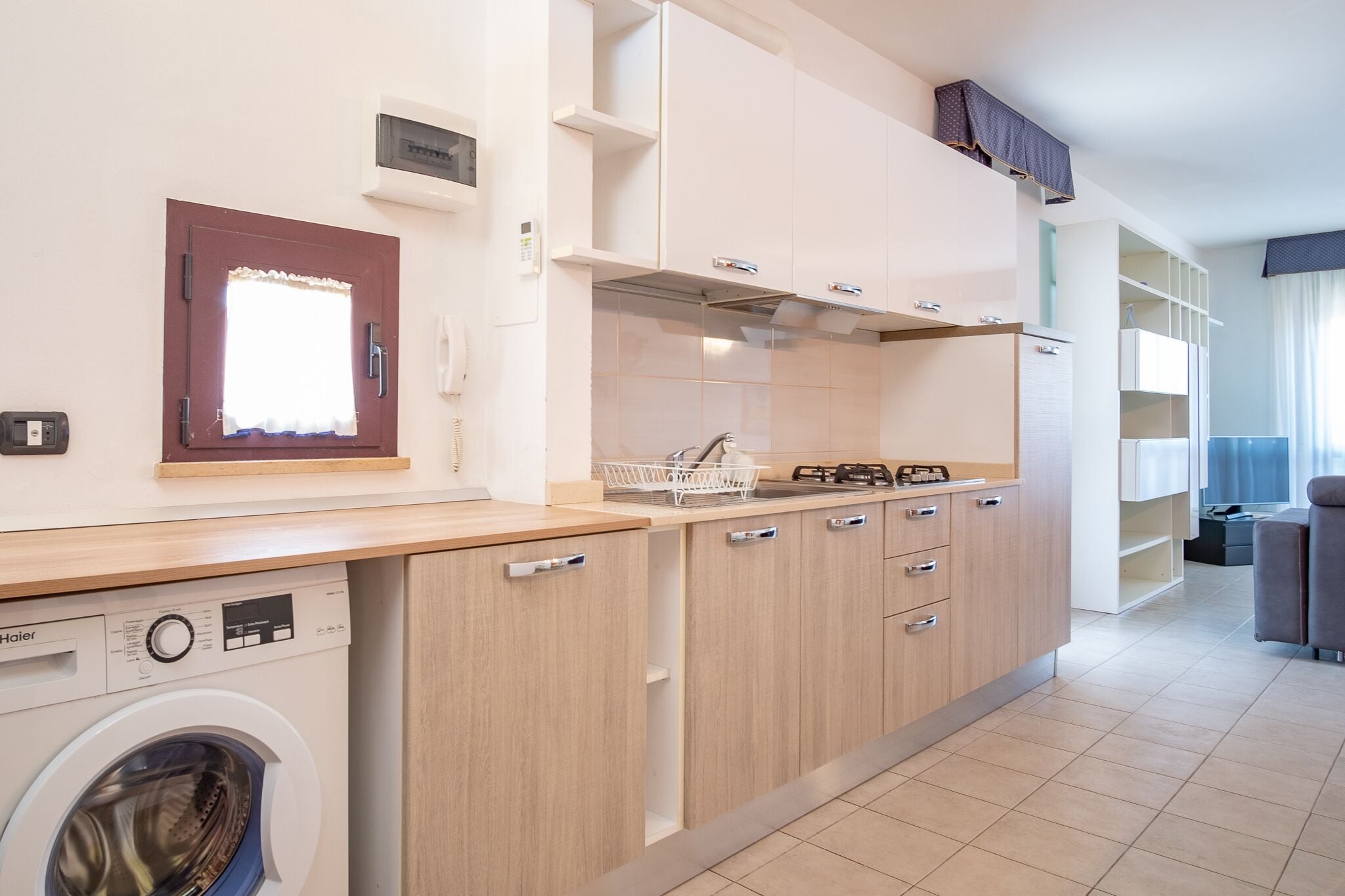 A compact kitchen with a built-in washer, wooden cabinets, and a window with curtains.