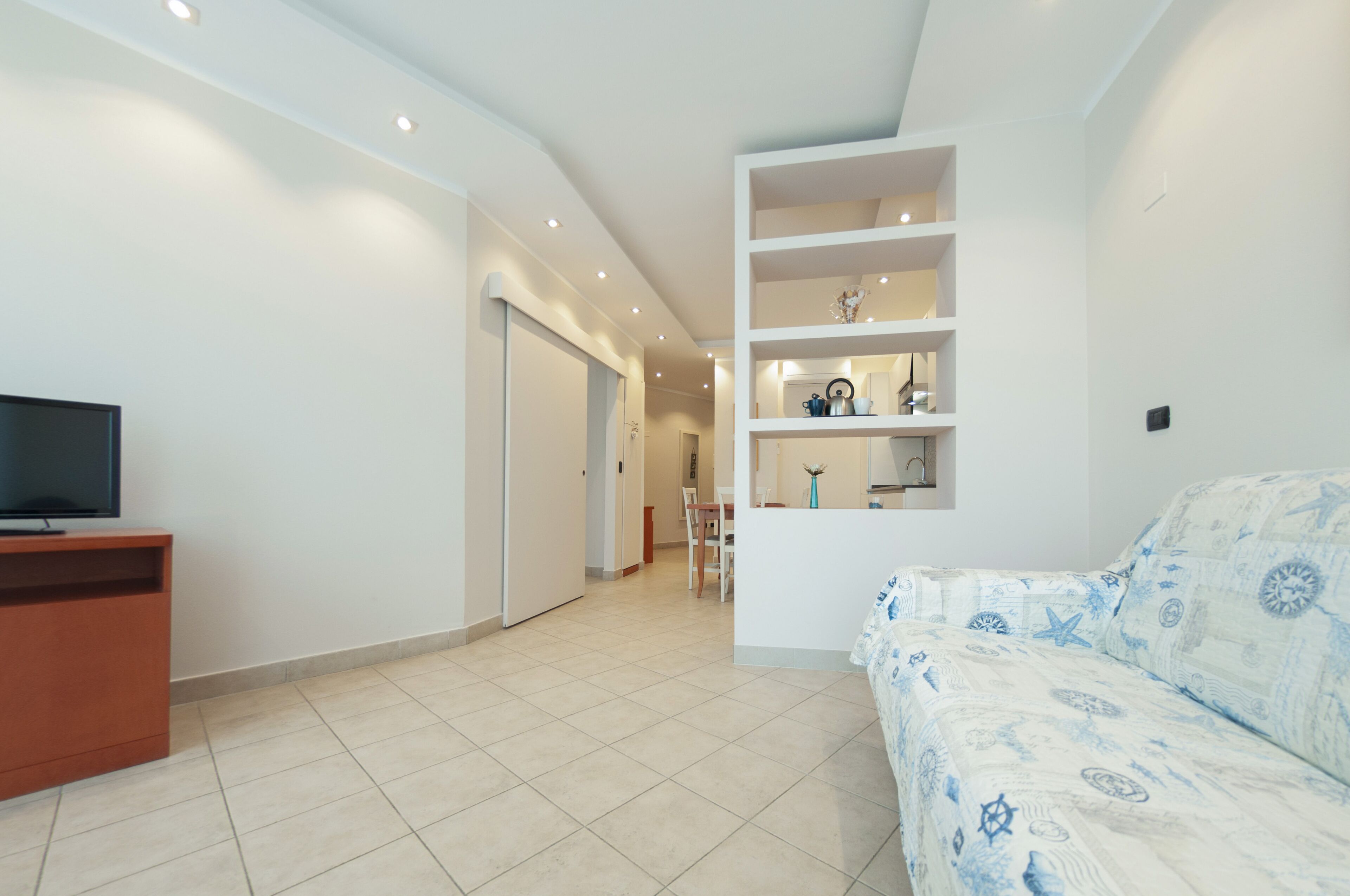 A modern hotel room with a flat-screen TV, a sofa with a blue and white pattern, a built-in shelving unit, and a tiled floor.